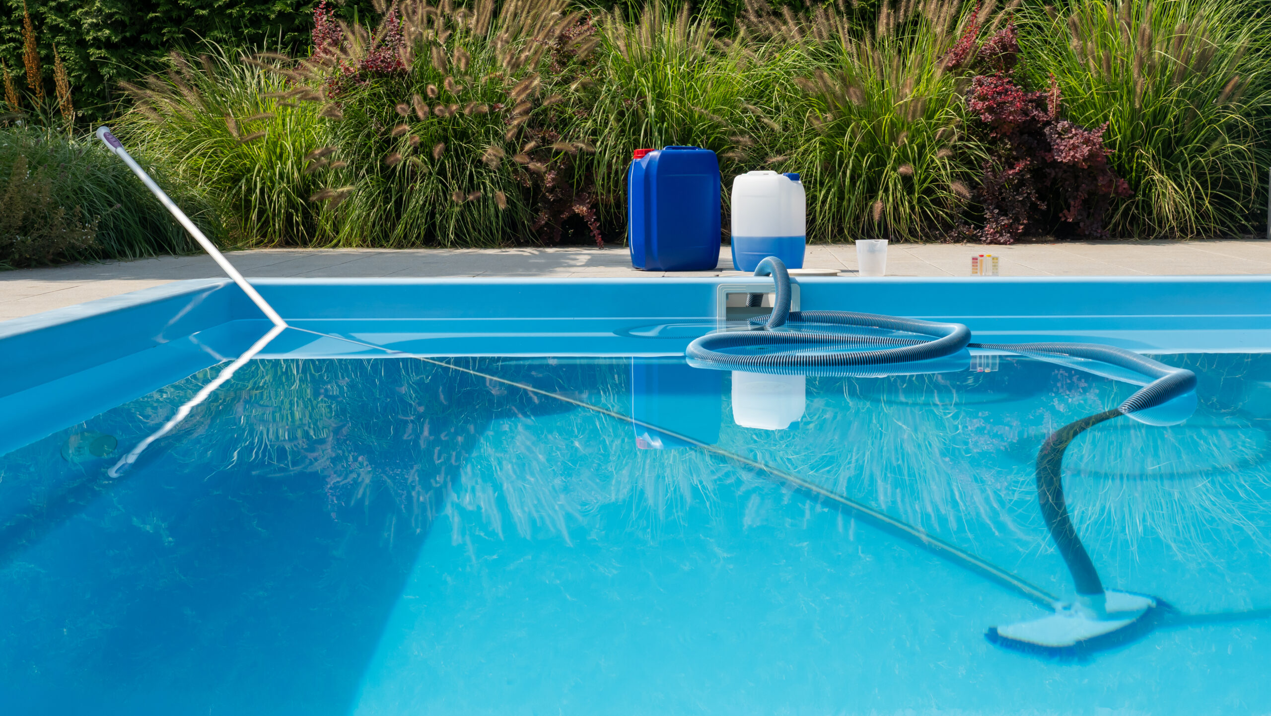 a clear blue pool with a submerged cleaning hose, chemical containers, and a water testing kit on the poolside, framed by lush greenery.