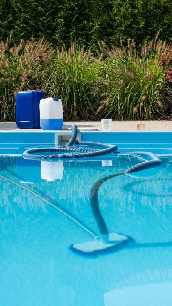 pool cleaning hose submerged in a clear blue pool, with chemical containers and testing kit on the poolside, surrounded by lush greenery.